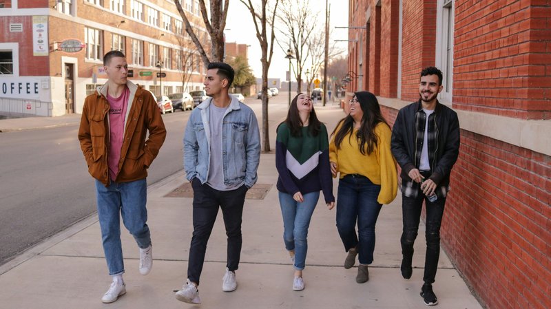 A group of five teenagers walk down the sidewalk next to a red brick building.