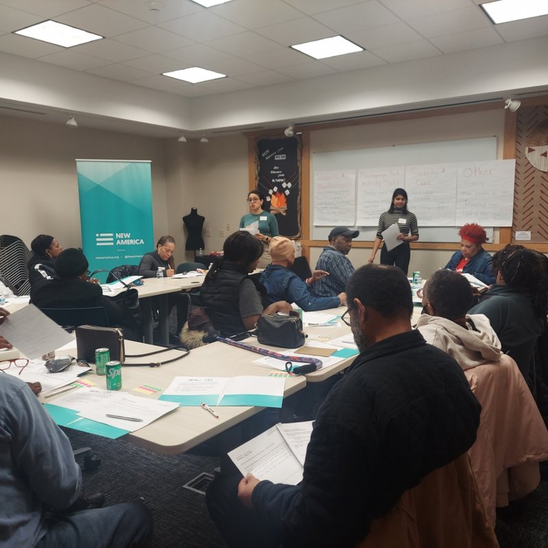 A group of people seated around tables in a bright room, engaged in a meeting.