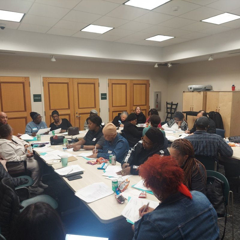 A group of people seated around tables in a bright room, engaged in a meeting.