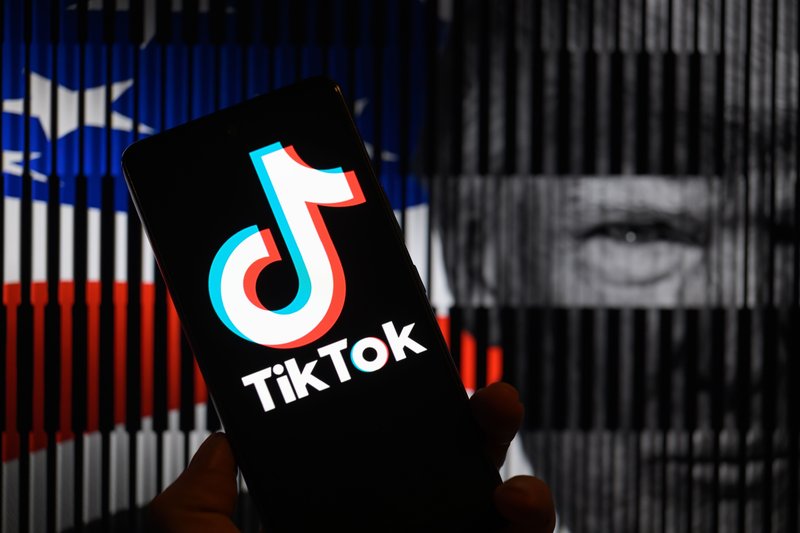 TikTok logo is being displayed on a mobile screen, in front of a photo illustration featuring the American flag and Trump.